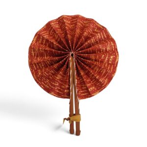 Éventail artisanal rond en tissu rouge brique et motifs dorés, avec manches en simili cuir marron attachés par un ruban.